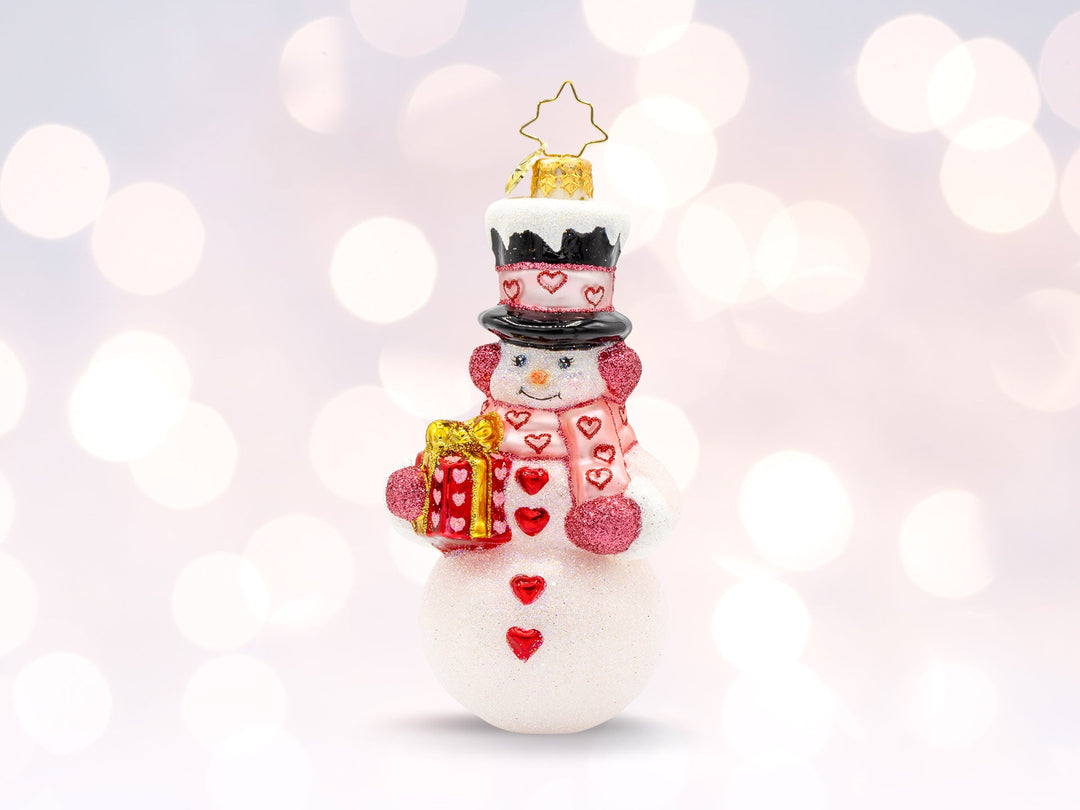 A Valentine's Day snowman ornament with red heart buttons, a red gift, a pink heart scarf, and a black hat with pink heart ribbon.