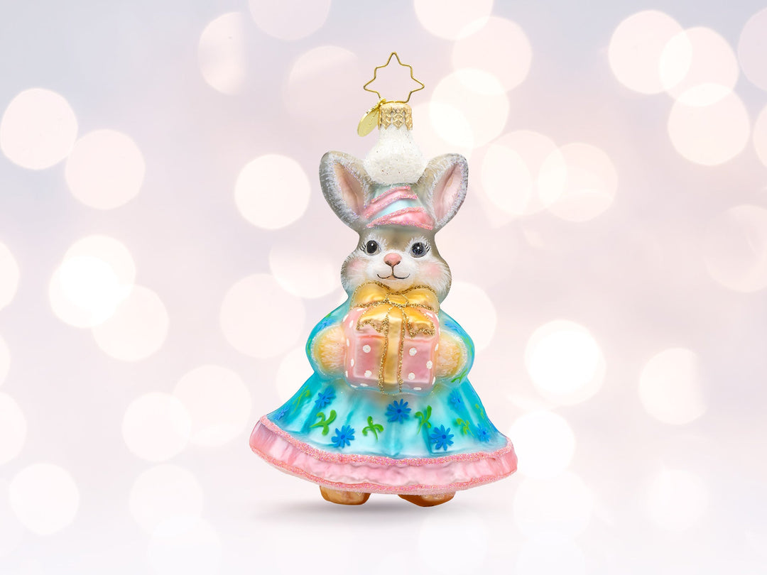 A grey easter bunny ornament wearing a blue and pink dress and hat, holding a pink gift.
