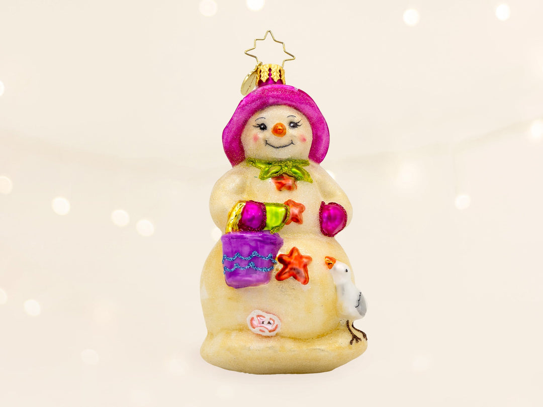 A sandy snowman ornament, wearing a pink beach hat, holding a purple sand bucket, and has a seagull on the side.