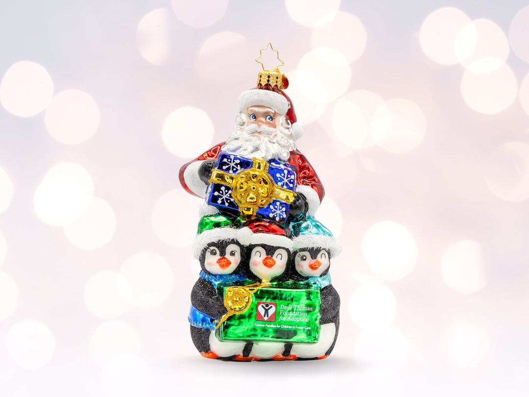 A classic red santa ornament holding a blue gift standing over three penugins holding a Dave Thomas Foundation for adoption sign.