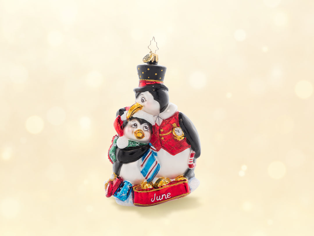 A father penguin ornament hugging a child penguin holding a tie. Below the penguins features the word June to celebrate Father's day.