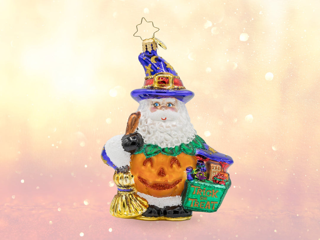 A Halloween santa ornament, wearing an orange pumpkin suit, and a purple wizard's hat, while holding a broom in one hand and a green trick or treat basket in the other.