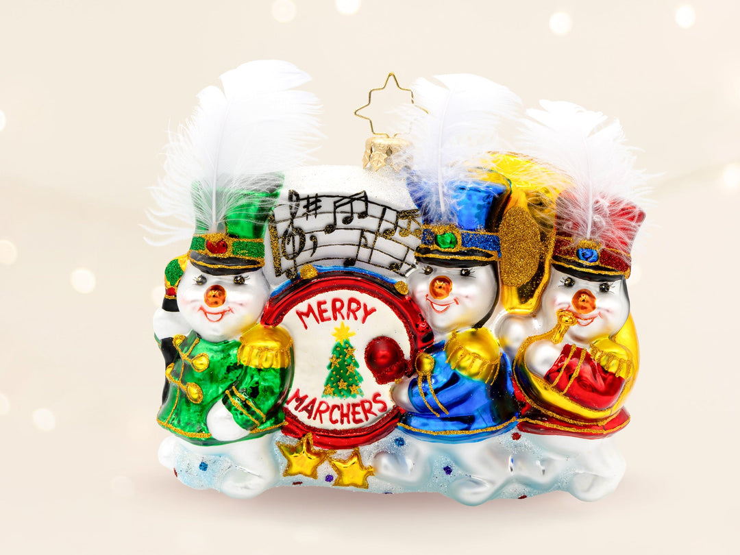 A snowman marching band ornament, with a snowman in green leading with a staff in front, followed by a snowman in blue pounding a Merry Marchers drum, and followed by a snowman in red playing a tuba.