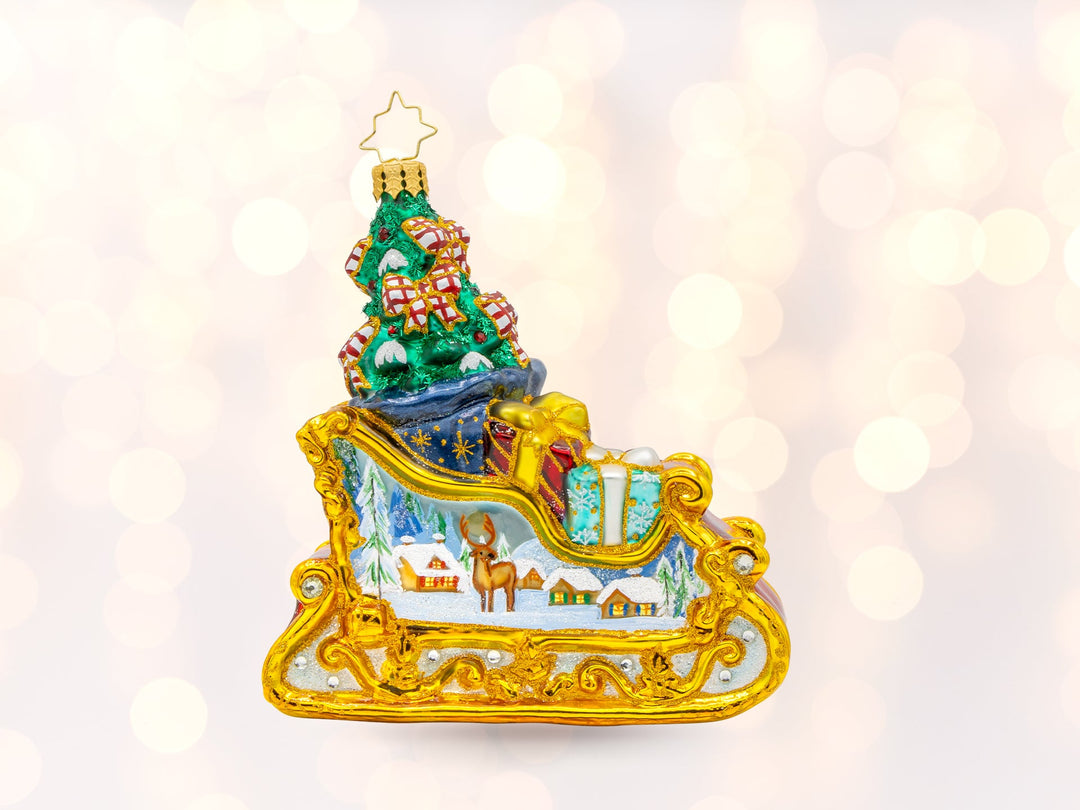 A gold sleigh ornament with a painted snowy cabin scene on the side of the sleigh. Inside of the sleigh is a pile of gifts and a Chrstimas tree in the back.
