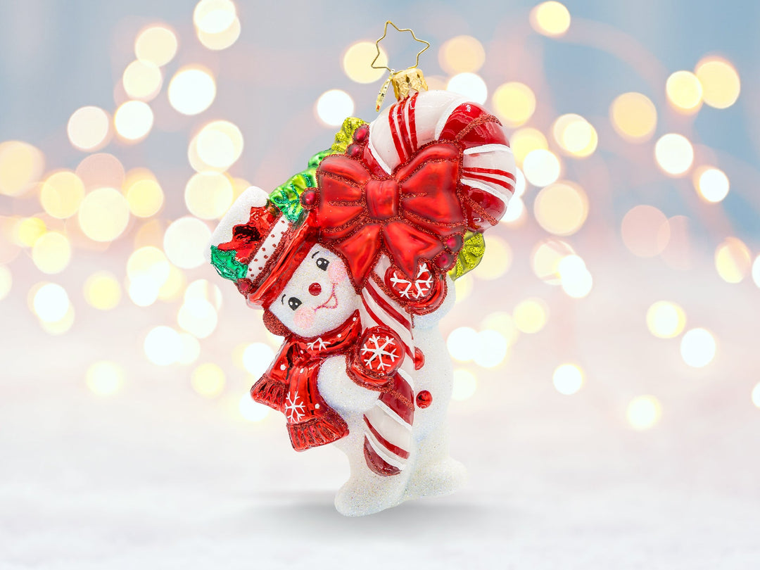 A snowman ornament wearing a red hat and scarf holding onto a large candy cane.
