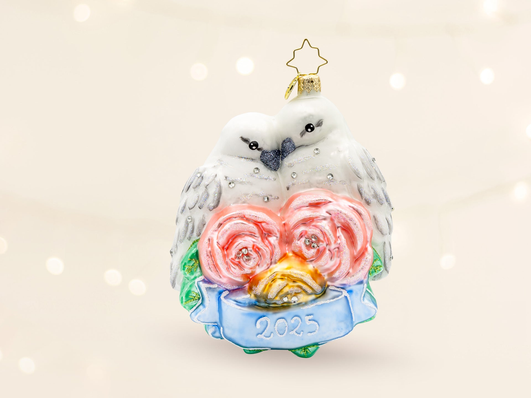 An ornament with a pair of white wedding birds nestled together with pink flowers and the year 2025 marked below on a blue ribbon.