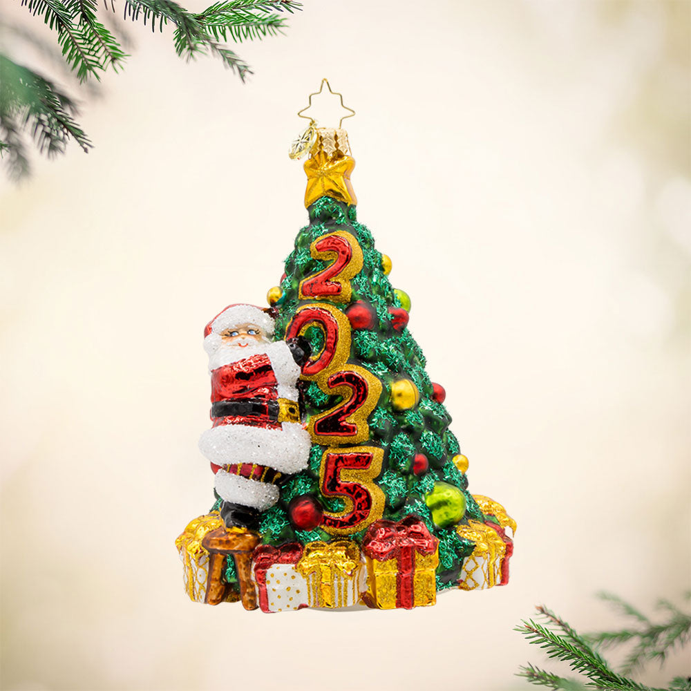 Santa's 2025 Tree Trim. Santa is seen decorating the Christmas tree with all the trimmings. Tree is dated with "2025." View of front.