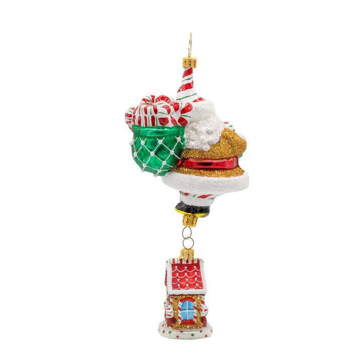 Ornament features a gingerbread house dangling from Santa's feet. Santa is holding a bag overflowing with treats. View of back.