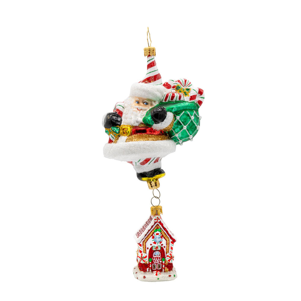 Ornament features a gingerbread house dangling from Santa's feet. Santa is holding a bag overflowing with treats. View of front.
