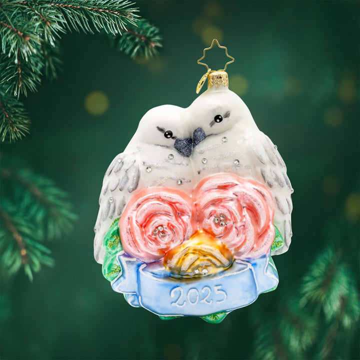 Wedding Lovebirds ornament. White birds are nestled into one another accompanied by pink flowers and a blue banner dated "2025." View of front.