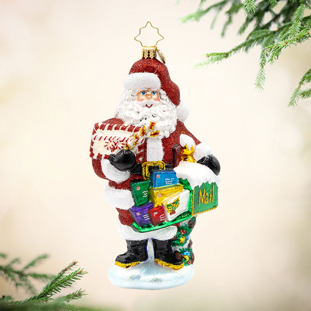 North Pole Mail Delivery ornament. Santa dons his red suit, standing next to a green mailbox filled with letters. View of front.