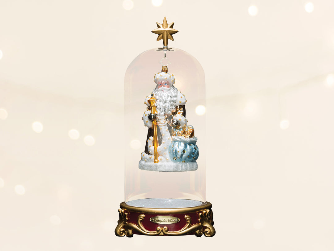 A blue and white robed santa ornament holding a gold cane and a blue bag. The ornament is suspended withing a glass dome display topped with a gold star.