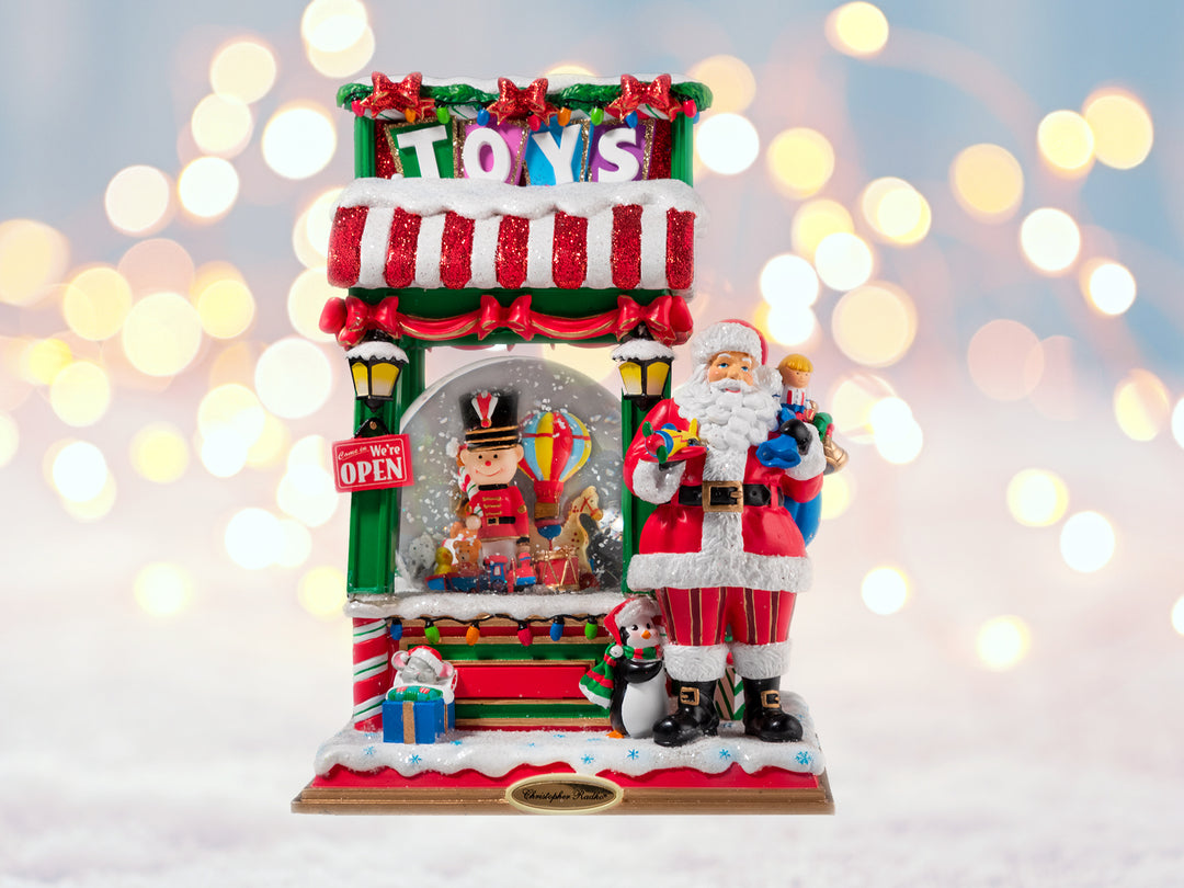 A santa toy shop snowglobe. Santa is standing outside the toyshop holding a bag of toys. Inside the toyshop is a snowglobe featuring a toy soldier, animal train, and hot air balloon.