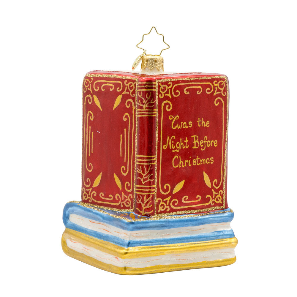 Ornament features Santa Claus, the jolly old elf, engrossed in a classic holiday tale. Dressed in his traditional red suit and hat, Santa sits comfortably in a plush armchair, surrounded by stacks of books, including the beloved "Twas the Night Before Christmas. View of back.