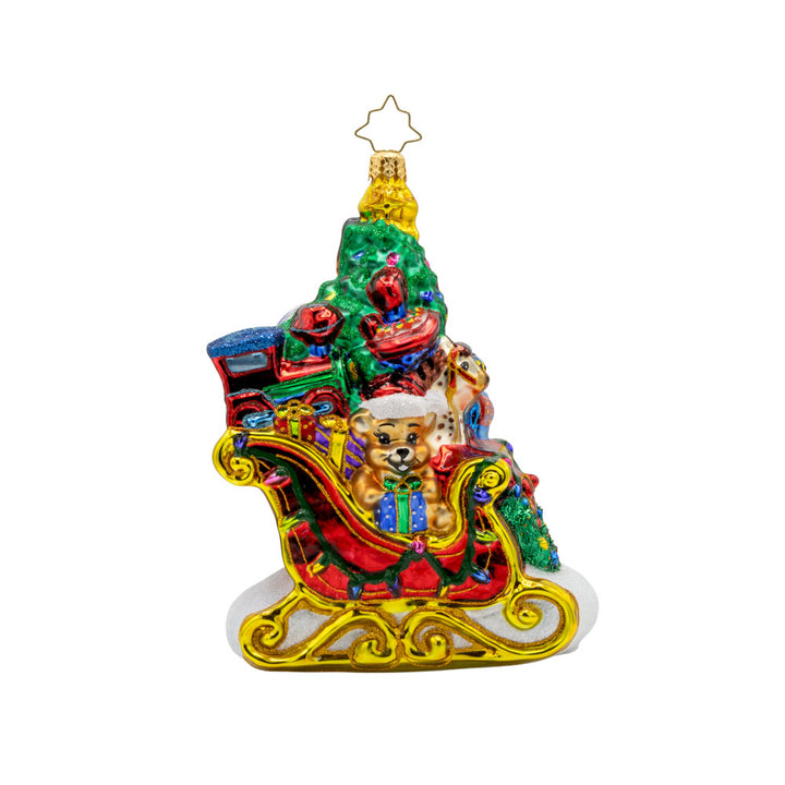 Jolly Sleigh Express ornament. A teddy bear, toy soldier, doll house, drum set, robot, rocking horse, and train engine are seen amidst the pile of presents in the sleigh. A Christmas tree is included in this festive scene. View of front.