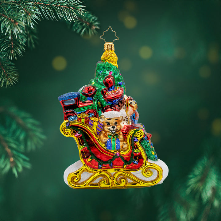 Jolly Sleigh Express ornament. A teddy bear, toy soldier, doll house, drum set, robot, rocking horse, and train engine are seen amidst the pile of presents in the sleigh. A Christmas tree is included in this festive scene. View of front.