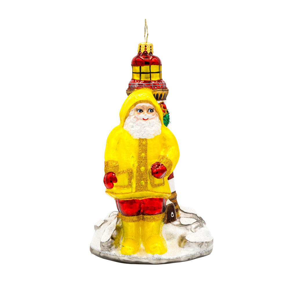 Santa's Coastal Haven ornament. Santa is wearing a yellow raincoat and boots standing next to a lighthouse. View of front.