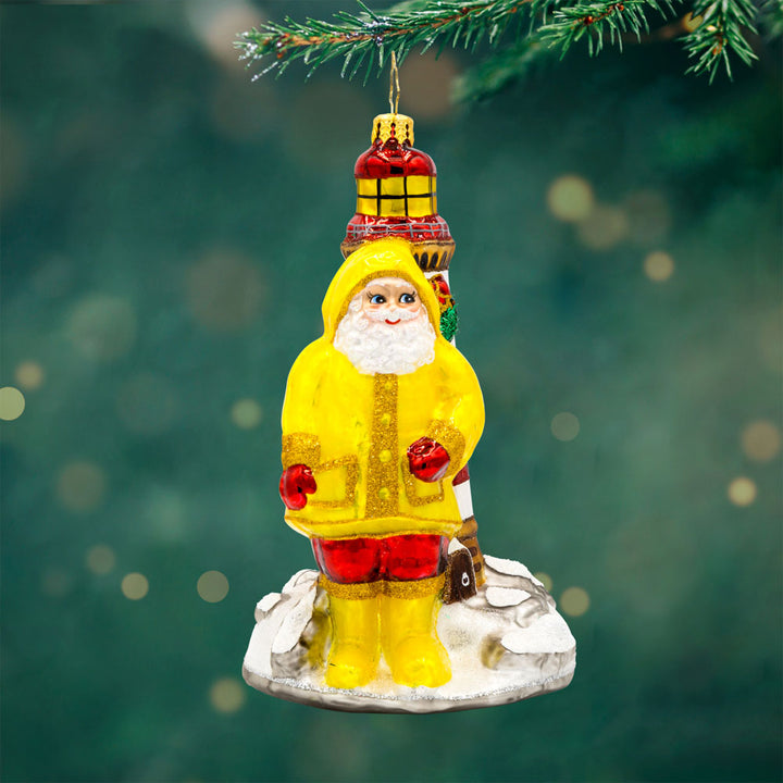 Santa's Coastal Haven ornament. Santa is wearing a yellow raincoat and boots standing next to a lighthouse. View of front.