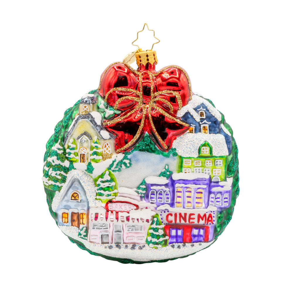 Christmas Village Charm ornament. Charming houses, twinkling lights, and a snowy scene topped with a red bow. View of front. 