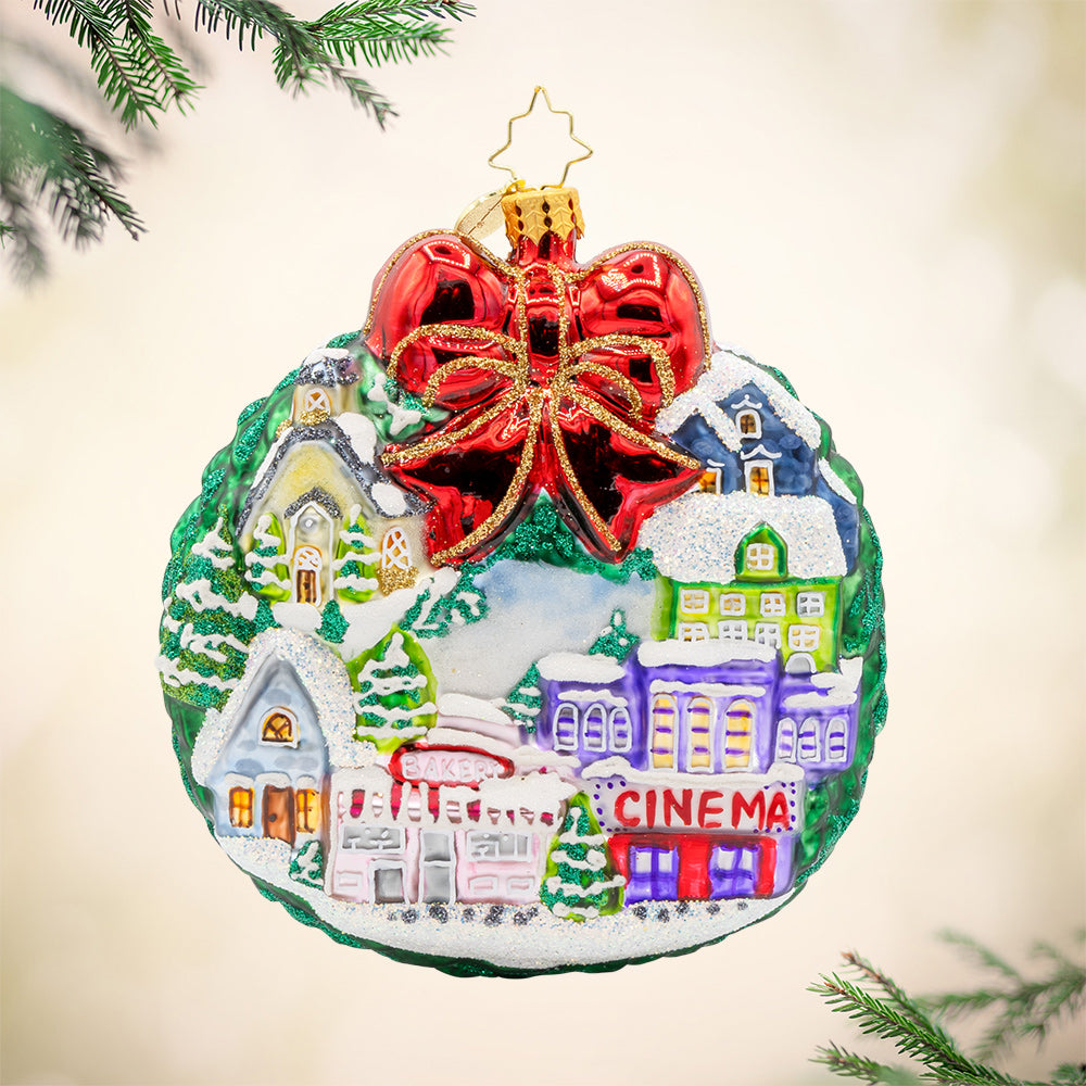 Christmas Village Charm ornament. Charming houses, twinkling lights, and a snowy scene topped with a red bow. View of front. 