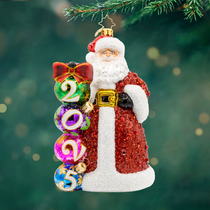 2025 Christmas Countdown ornament. Santa dons a glittering red jeweled coat.  Ornaments with "2025" written on them are stacked next to his side. View of front.
