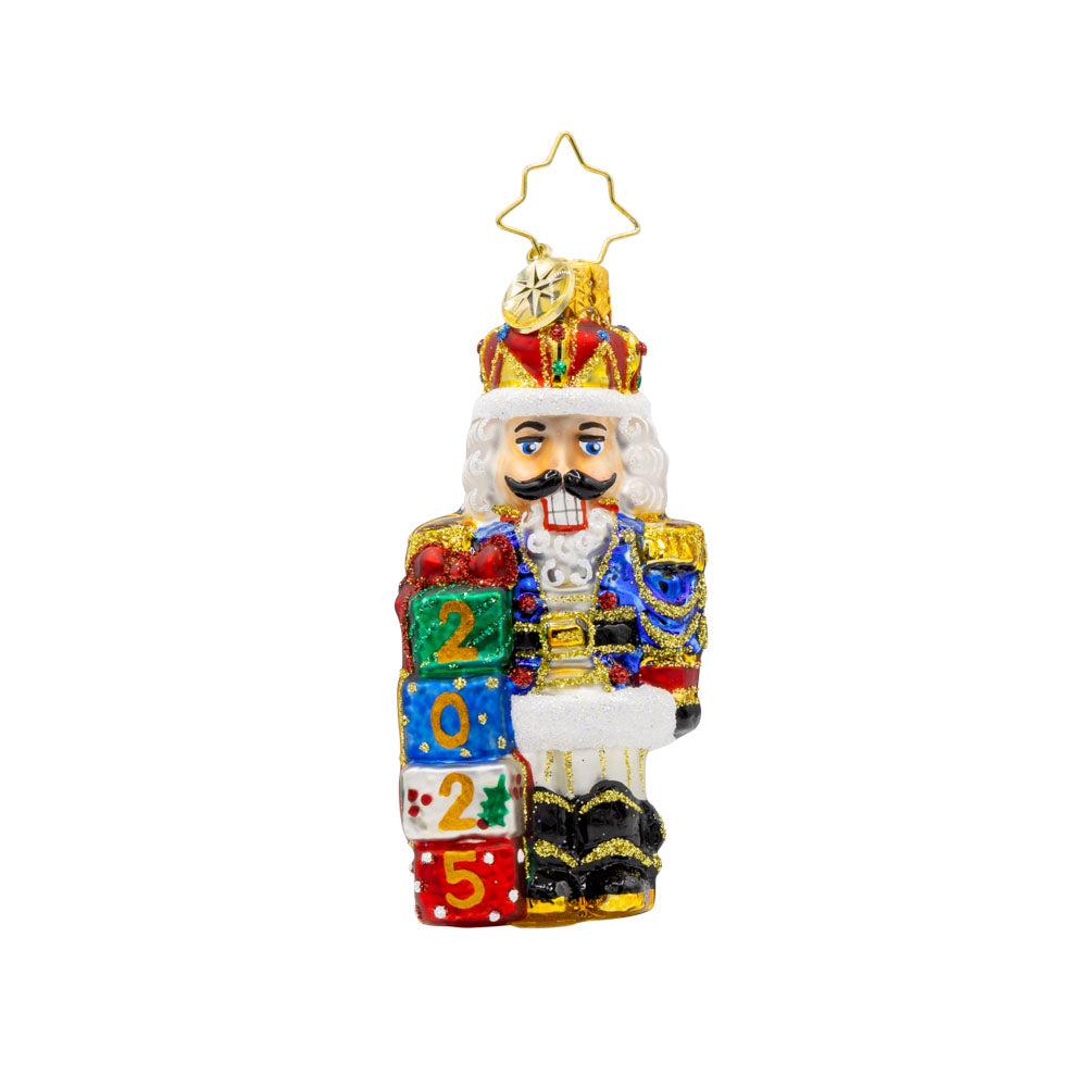 Royal Gift Guardian 2025 Gem ornament. This nutcracker dons a suit of blue, red, and gold, standing next to stacked gifts dated "2025." View of front. 