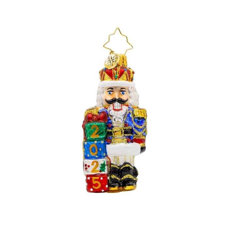 Royal Gift Guardian 2025 Gem ornament. This nutcracker dons a suit of blue, red, and gold, standing next to stacked gifts dated "2025." View of front. 