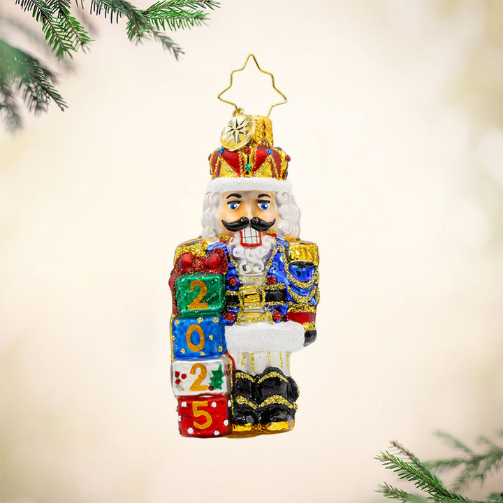 Royal Gift Guardian 2025 Gem ornament. This nutcracker dons a suit of blue, red, and gold, standing next to stacked gifts dated "2025." View of front. 