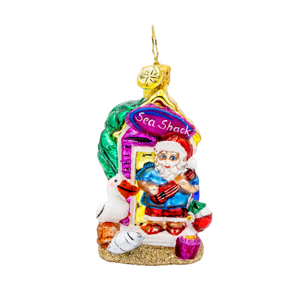 Santa's Seaside Shack Gem ornament.  Santa stands outside a sea shack ukulele in hand. A pelican and seashells keep him company. View of front. 