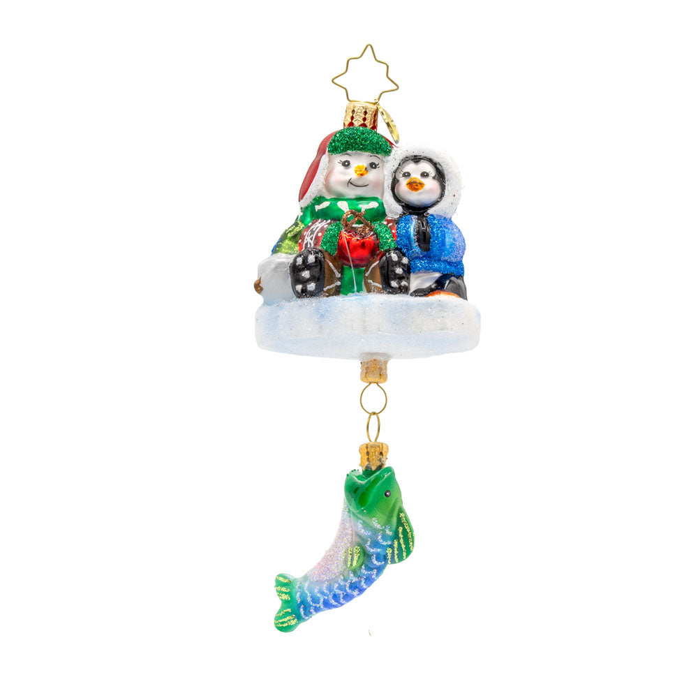 Ice Fishing Snow Pals Gem ornament. A snowman and penguin sit bundled up on a block of ice with a fishing pole. A fish dangles below. View of front. 