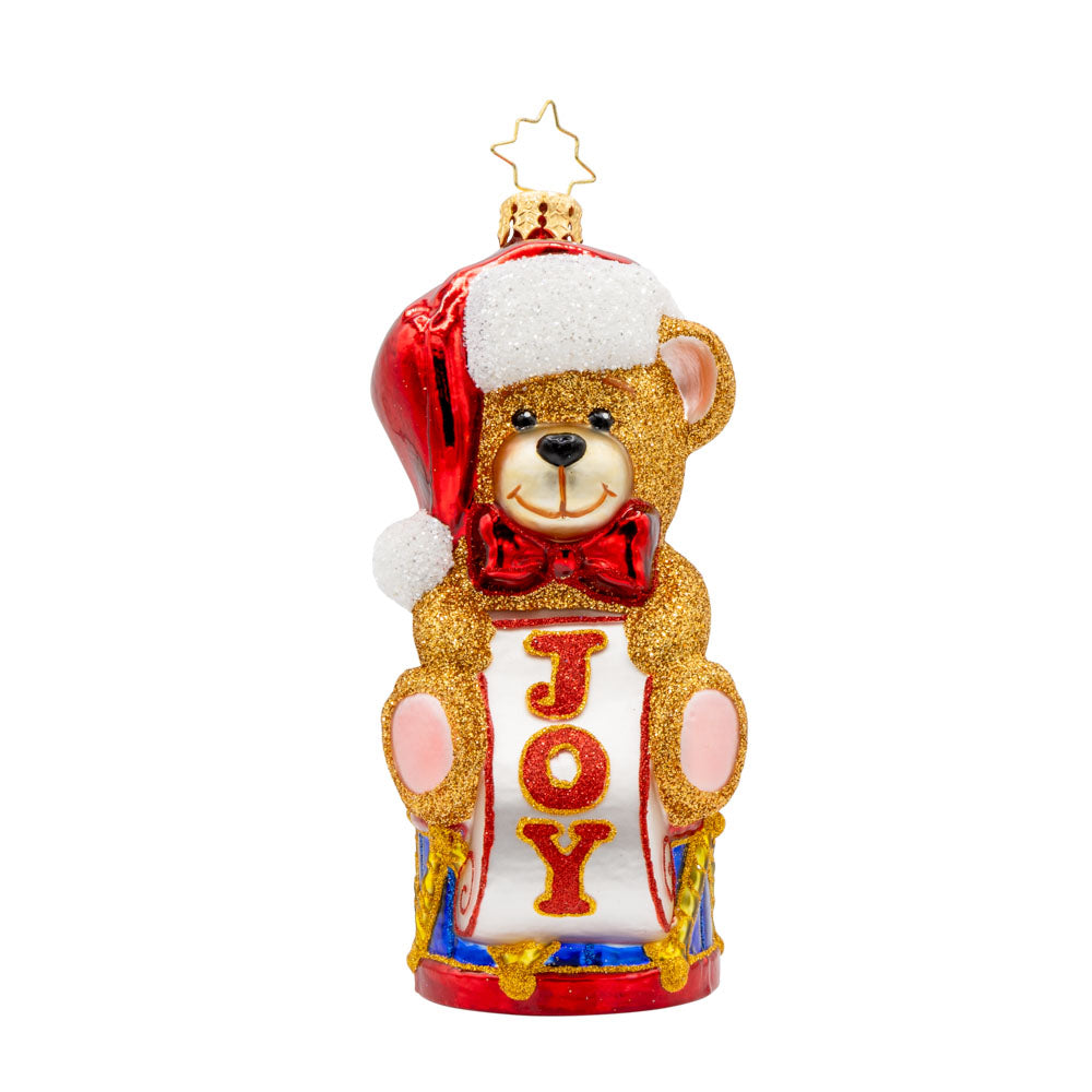 Teddy Bear Greetings ornament. Teddy bear wearing a Santa hat sitting atop a drum holding a sign that says "JOY." View of front. 