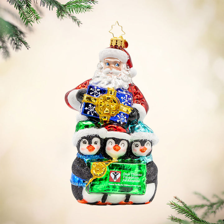Santa Flurry Friends ornament. Santa is standing with three penguins in front of him, holding a gift with the Dave Thomas Foundation for Adoption logo on the front of it. View of front. 