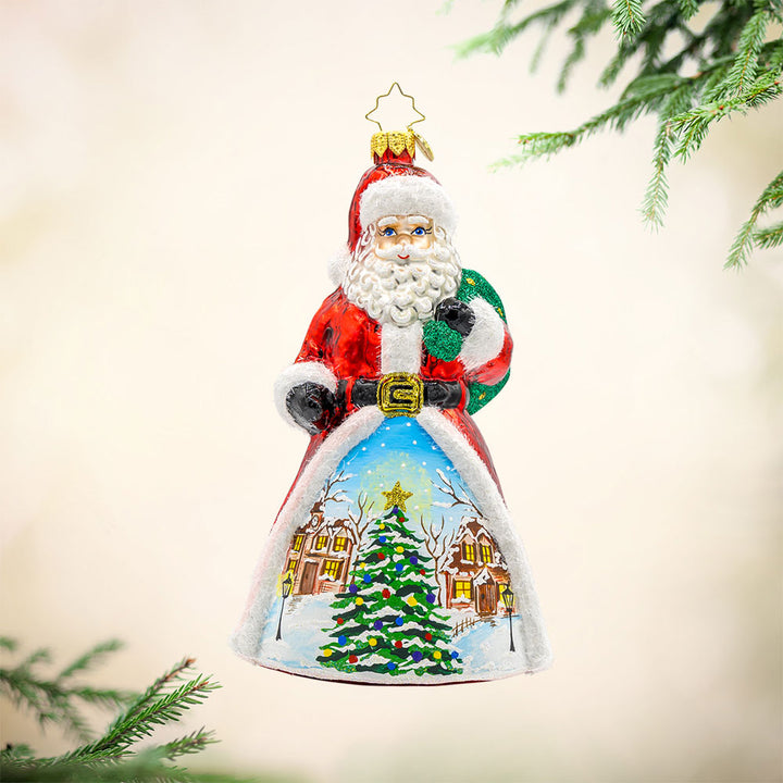 St. Nick's Winter Wonderland ornament. Santa stands amidst a picturesque snowy village. The hand-painted scene features twinkling lights and cozy homes surrounding a Christmas tree in the center. View of front.
