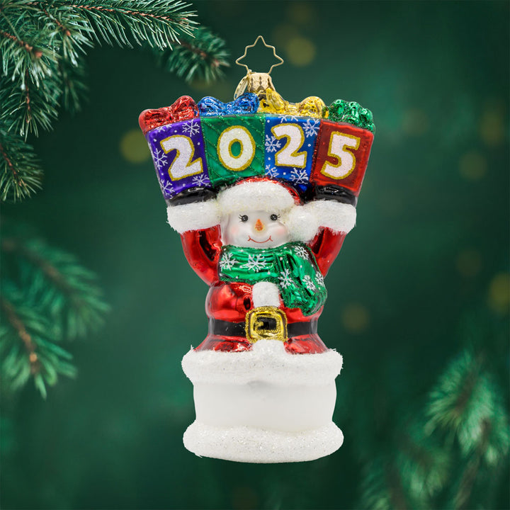 2025 Snowman Greetings ornament. A snowman dressed as Santa raises a stack of gifts dated "2025" above his head. View of front. 