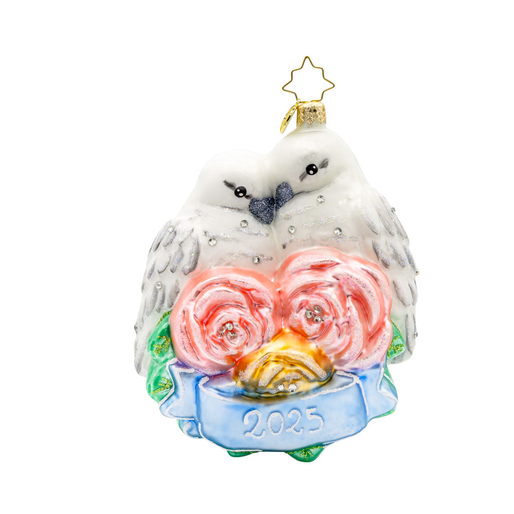 Wedding Lovebirds ornament. White birds are nestled into one another accompanied by pink flowers and a blue banner dated "2025." View of front. 