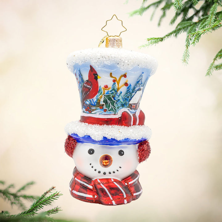 Snowy and the Cardinals ornament. This snowman ornament features a hand-painted winter scene, complete with a cozy cabin, a vibrant cardinal, and a snowy landscape. He wears a festive hat, topped with a snowy peak, sparkling glitter muffs and satin scarf. View of front. 