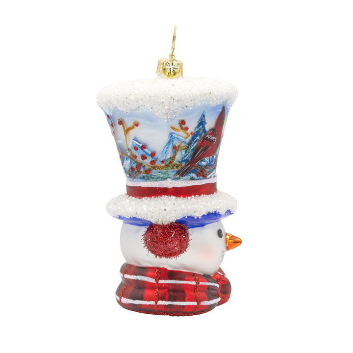 Snowy and the Cardinals ornament. This snowman ornament features a hand-painted winter scene, complete with a cozy cabin, a vibrant cardinal, and a snowy landscape. He wears a festive hat, topped with a snowy peak, sparkling glitter muffs and satin scarf. View of left side.