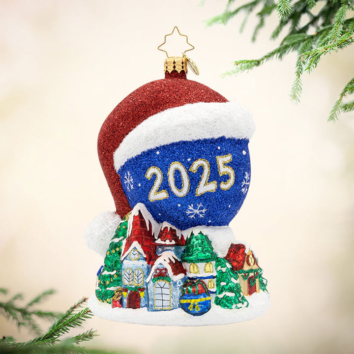 Twas the Night Before Moonlight ornament. Dressed in a Santa hat, this glowing moon dated with "2025" keeps a watchful eye on the Christmas village below. View of front.