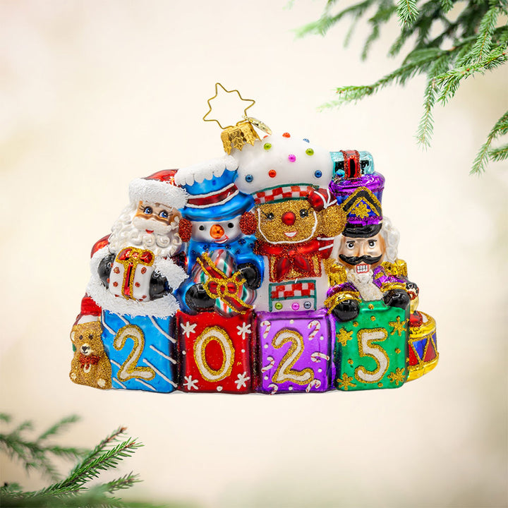 Stacking Up The Cheer ornament. A row of gifts dated with "2025." Behind them sits a snowman, gingerbread man, nutcracker and drummer boy. View of front. 