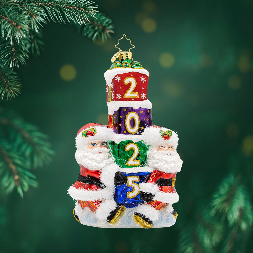 Gift Giving Santa's 2025 ornament. Two Santa's carry a stack of gifts with "2025" written on them. View of front. 