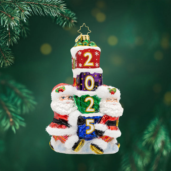 Gift Giving Santa's 2025 ornament. Two Santa's carry a stack of gifts with "2025" written on them. View of front. 