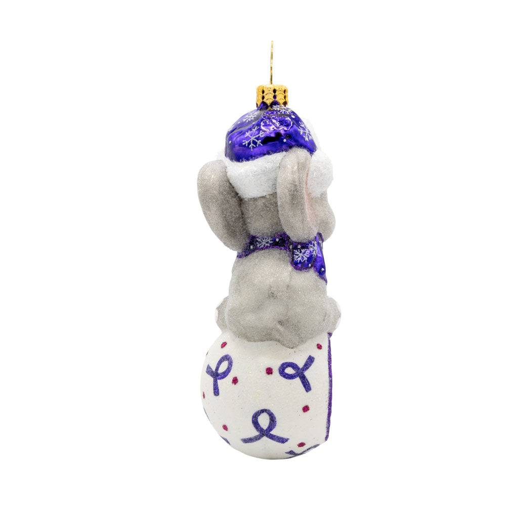 Trunk of Remembrance ornament. An elephant wears a purple scarf and hat sitting atop a white ball decorated with purple Alzheimer's awareness ribbons. View of side.