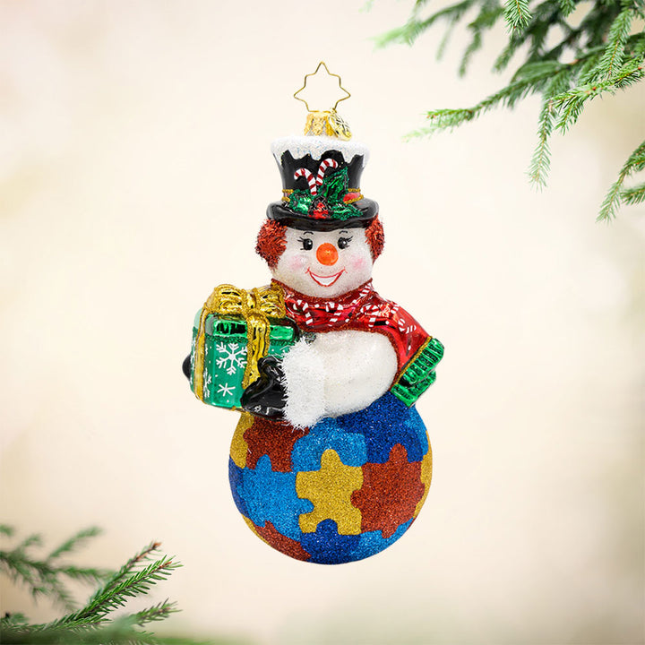 Puzzle Piece Pal ornament. This snowman is carrying a gift. His bottom half is painted to look like a jigsaw puzzle. View of front. 