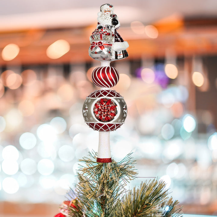 This enchanting finial features Santa, with his rosy cheeks, snowy beard, and a sack full of gifts above two decorated rounds. View of front displayed on tree.