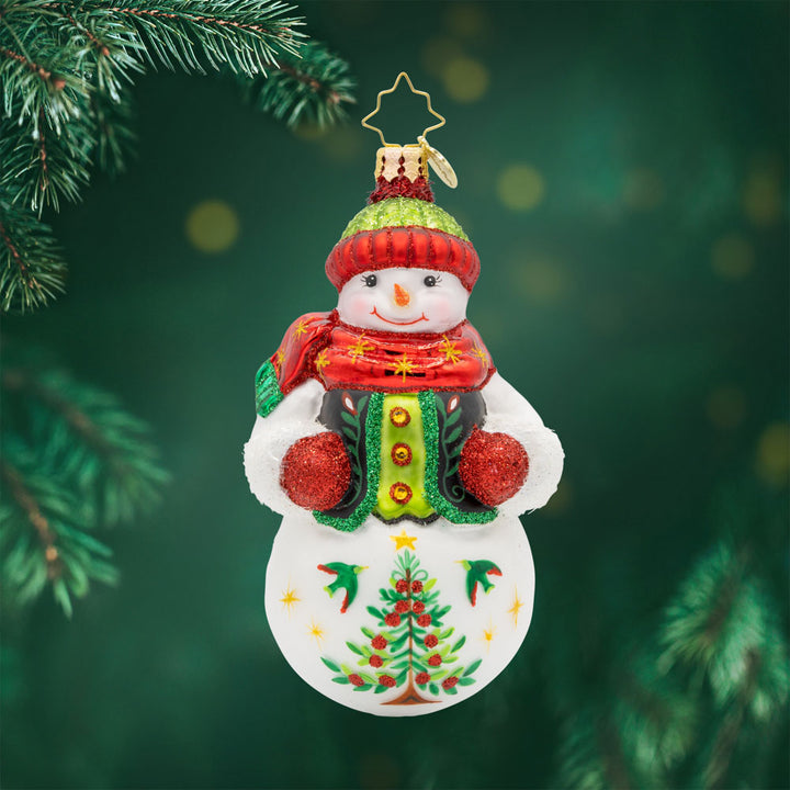 Festive Folk Flair ornament. Dressed in a cozy vest with hand-painted folk-art designs and a cheerful pom-pom hat. His glittering gloves and shining scarf bring the sparkle, while a Christmas tree adorned with stars and berries completes his festive look. View of front. 