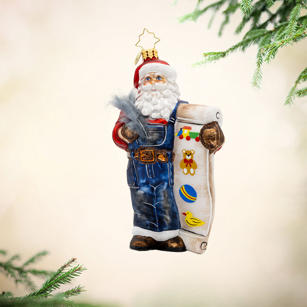 Santa's Holiday Roll Call ornament. Santa is wearing blue work overalls and is holding a long list of toys with quill in hand. View of front. 