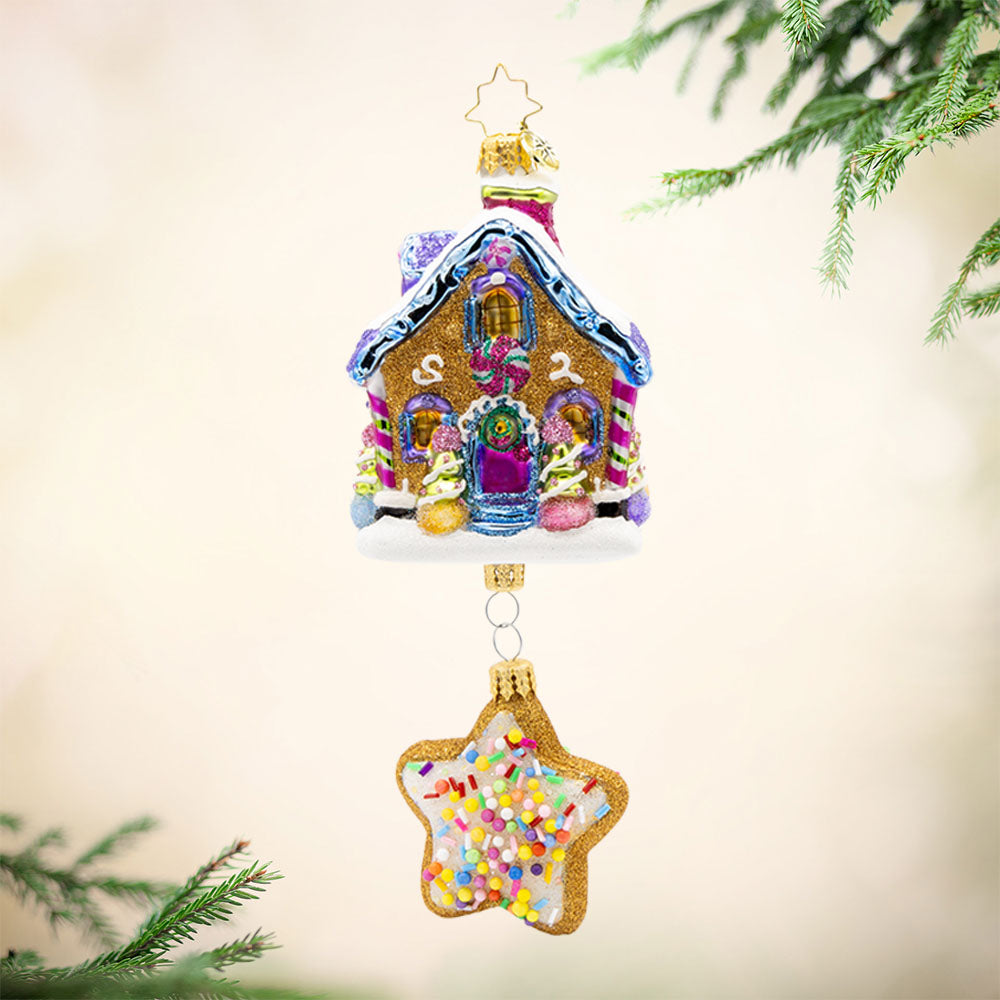 Sweet Beginnings ornament. A gingerbread house decorated in vibrant colors and candy. A gingerbread star cookie covered in sprinkles hangs below. View of front. 