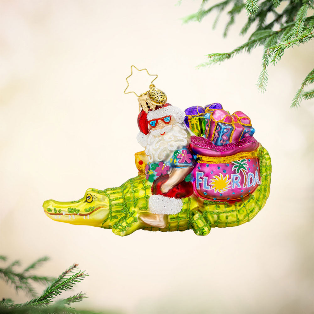 Sunshine State Gator ornament. Santa is trading in his sleigh for a gator and enjoying a tropical cocktail. "Florida" is written on gift sack. View of front. 