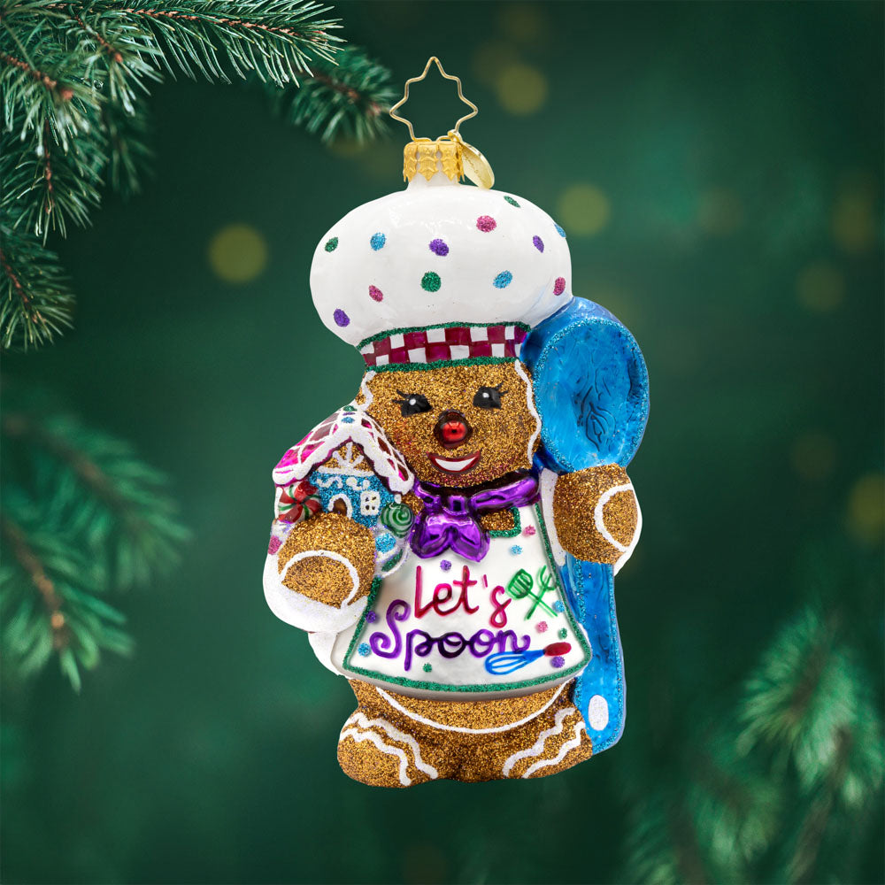Spoonful of Sweetness ornament. This gingerbread man is wearing a festive chef's outfit, spoon in hand, and gingerbread house in the other. View of front. 