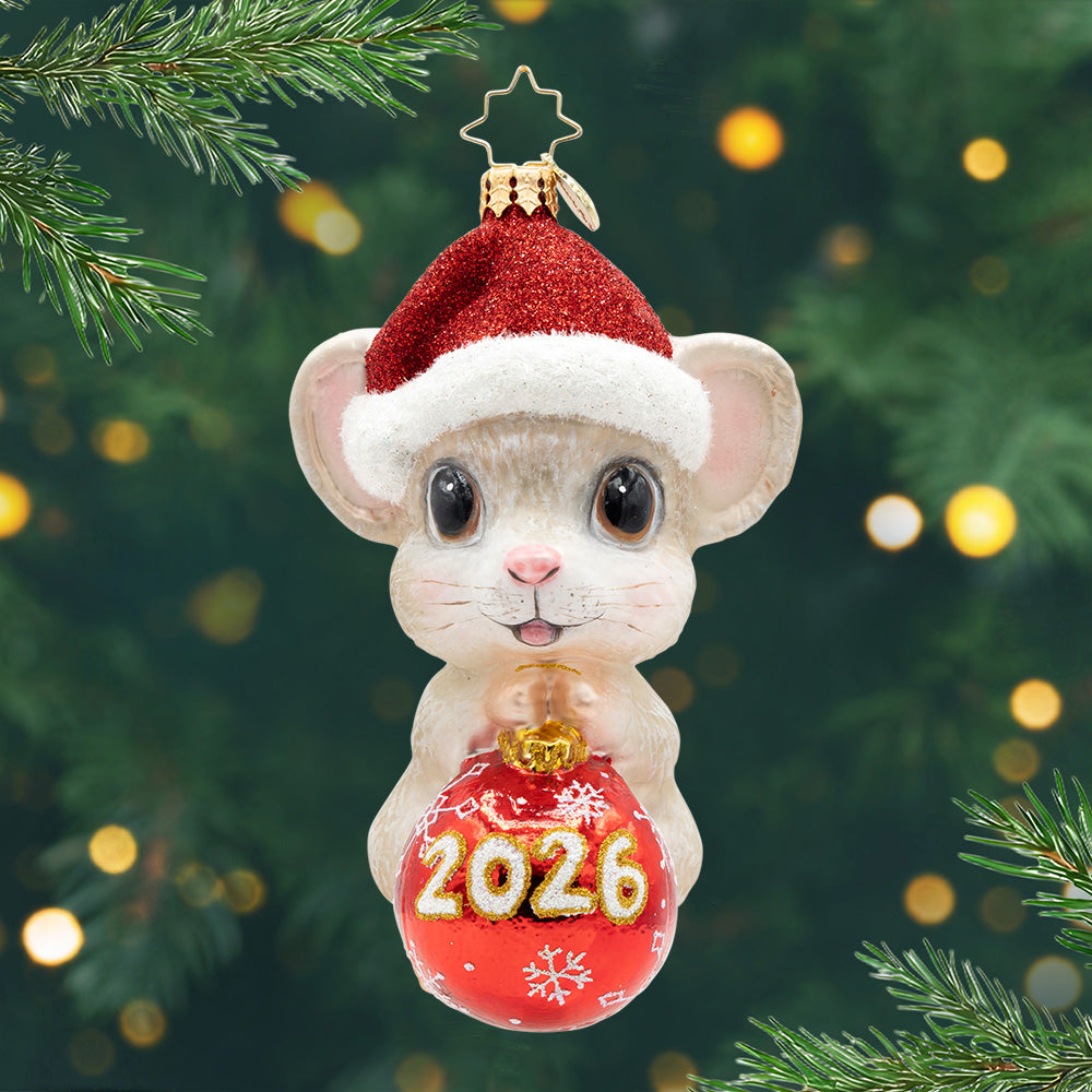2026 Tiny Timekeeper. Ornament of a light brown mouse wearing a Santa hat and holding a red ornament with the year 2026. View of front